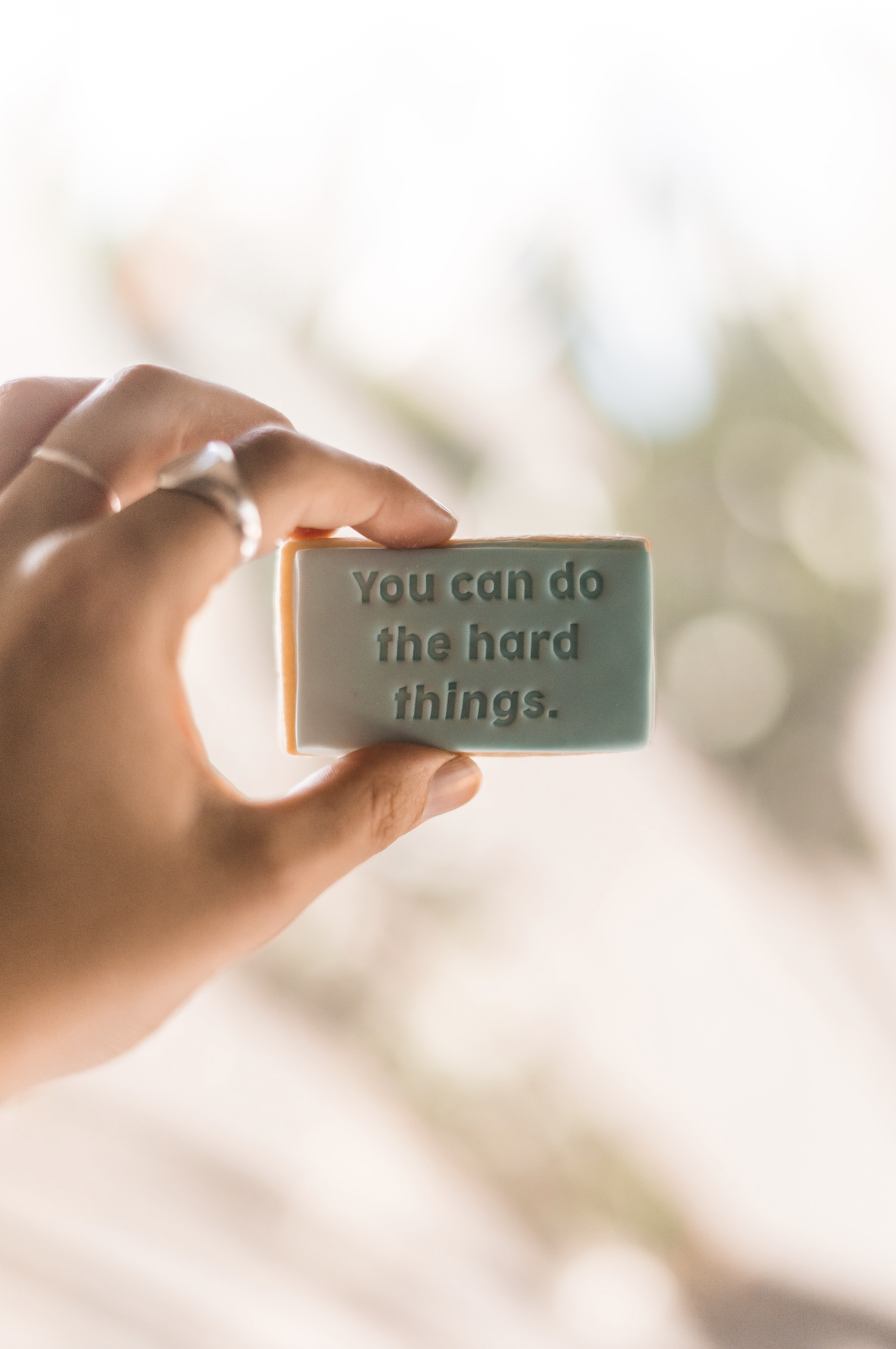 woman's hand holding up white sign in front of a window that says "you can do the hard things"