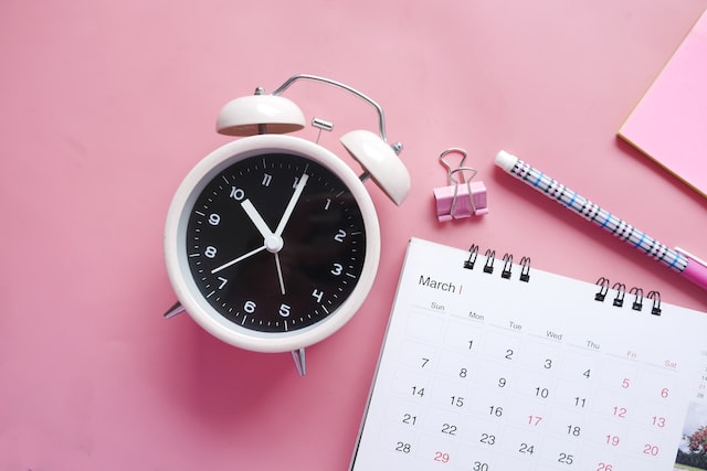 pink alarm clock next to a calendar and pencil on a pink background